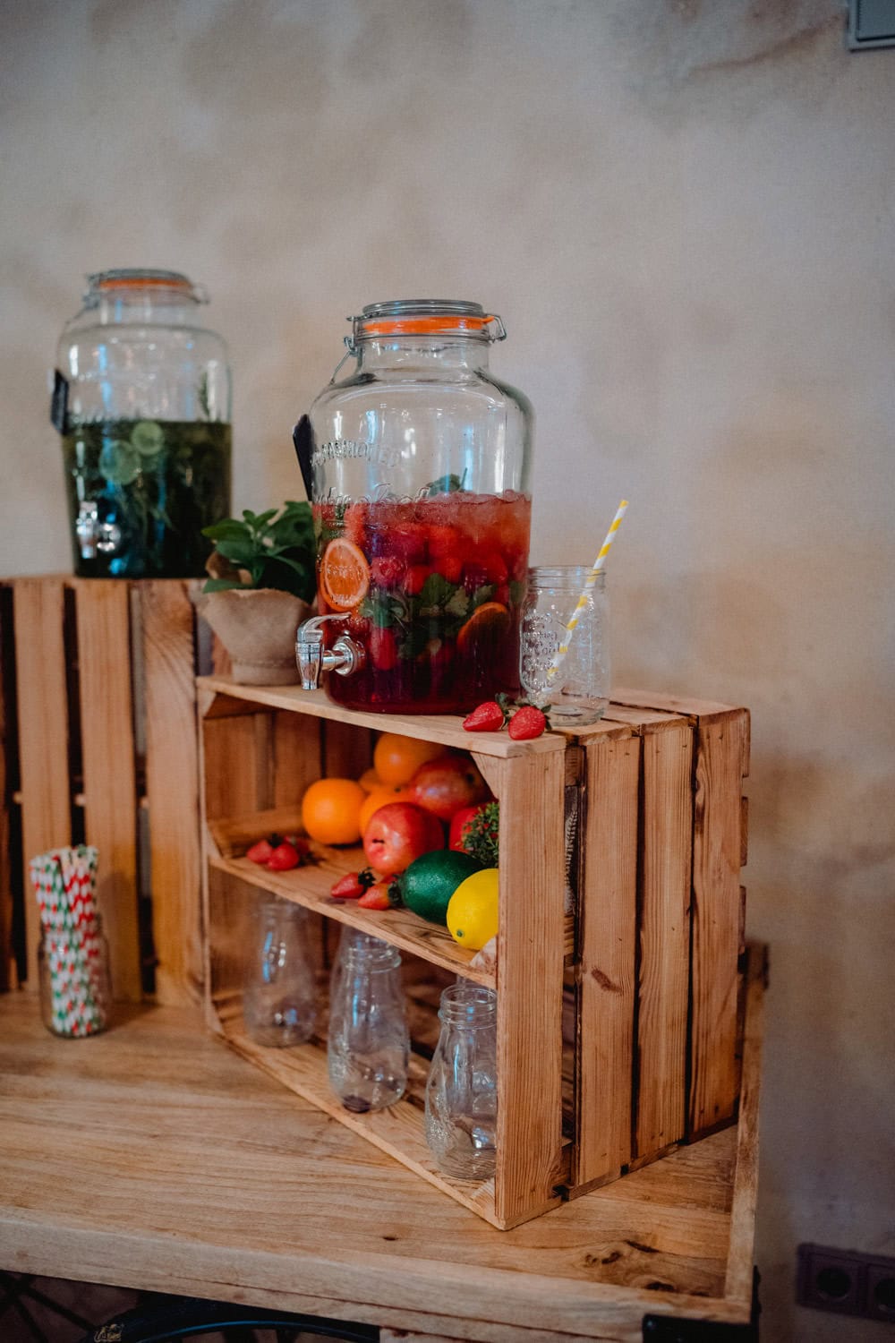 Im Hammerschloss Stodl Schmidmühlen stehen Getränkespender elegant auf einem Holzständer – einer ist randvoll mit roter Flüssigkeit und Fruchtscheiben, der andere mit einem grünen Getränk, garniert mit Minze. Darunter stehen leuchtende Orangen, Zitronen und Limetten neben leeren Gläsern und gestreiften Strohhalmen.