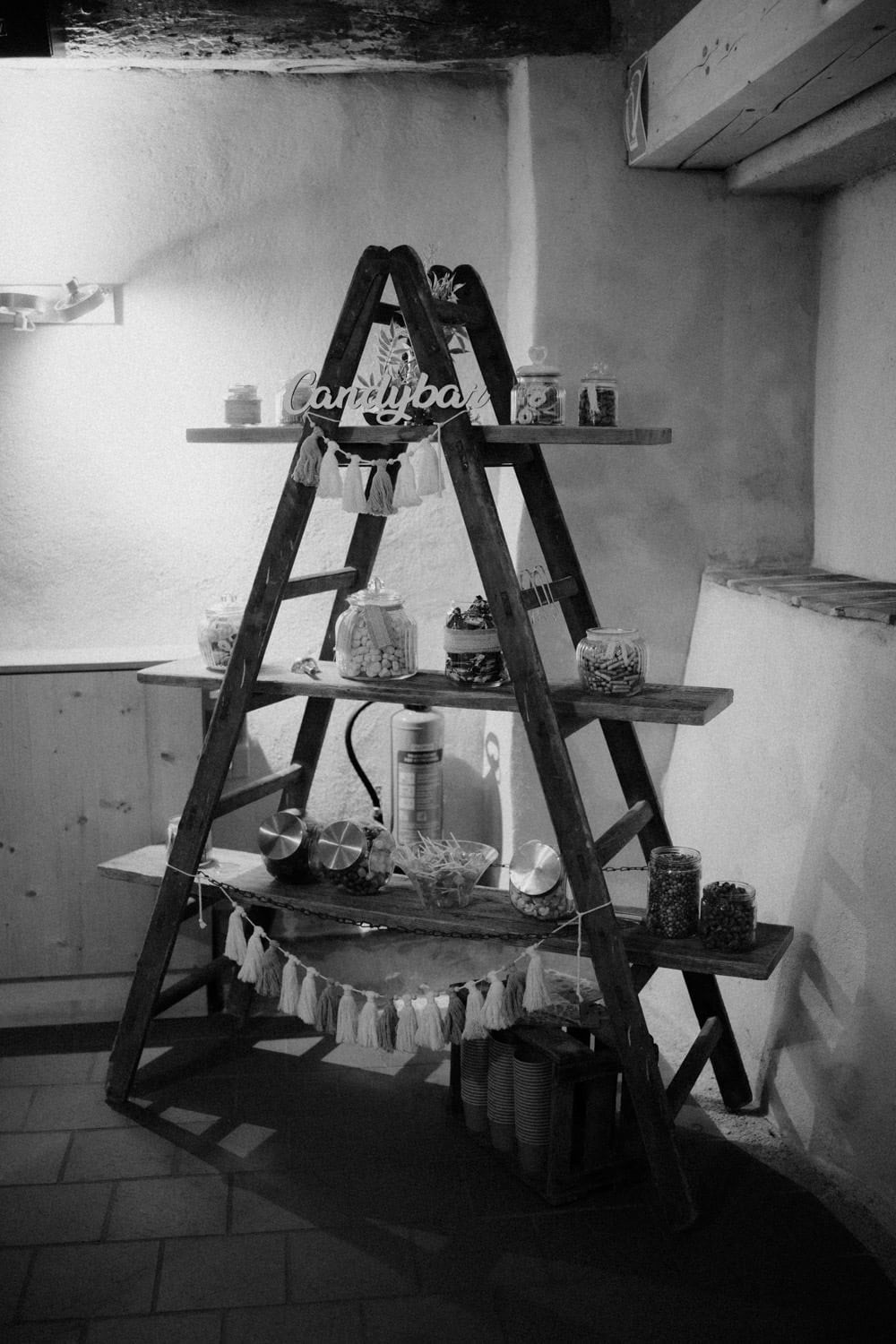 Ein Schwarzweißbild eines rustikalen hölzernen Leiterregals im Hammerschloss Stodl Schmidmühlen zeigt Gläser mit Bonbons, kleine Dekorationen und ein „Candybar“-Schild. Das Ganze ist elegant vor einer strukturierten Wand mit stimmungsvoller Beleuchtung angeordnet.