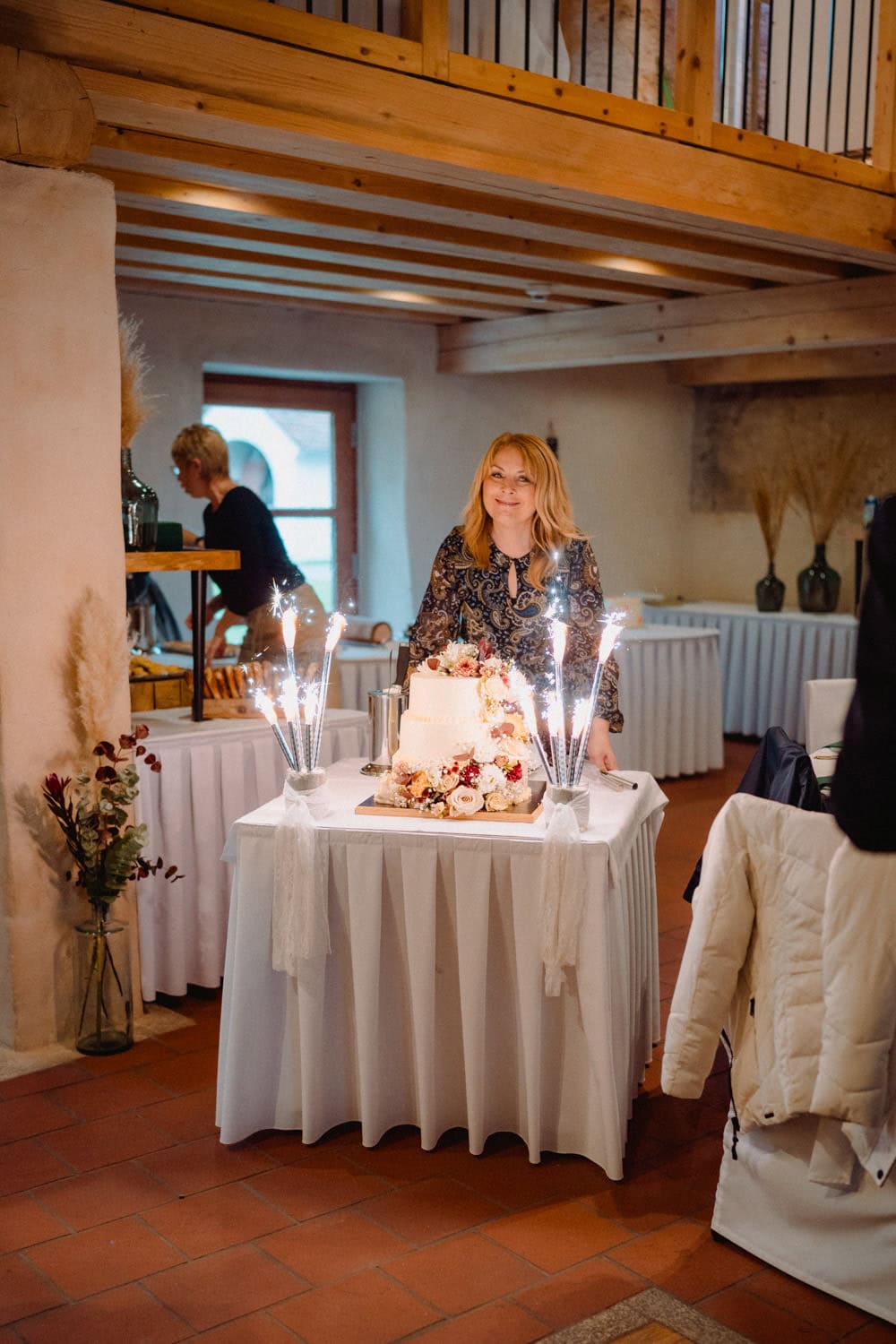 Eine Frau steht in einem rustikal eingerichteten Raum im Hammerschloss Stodl Schmidmühlen und lächelt neben einem dekorierten Tisch. Sie hält eine mehrstöckige Torte mit Blumen und Wunderkerzen in den Händen. Im Hintergrund sind Tische und Vasen elegant arrangiert.