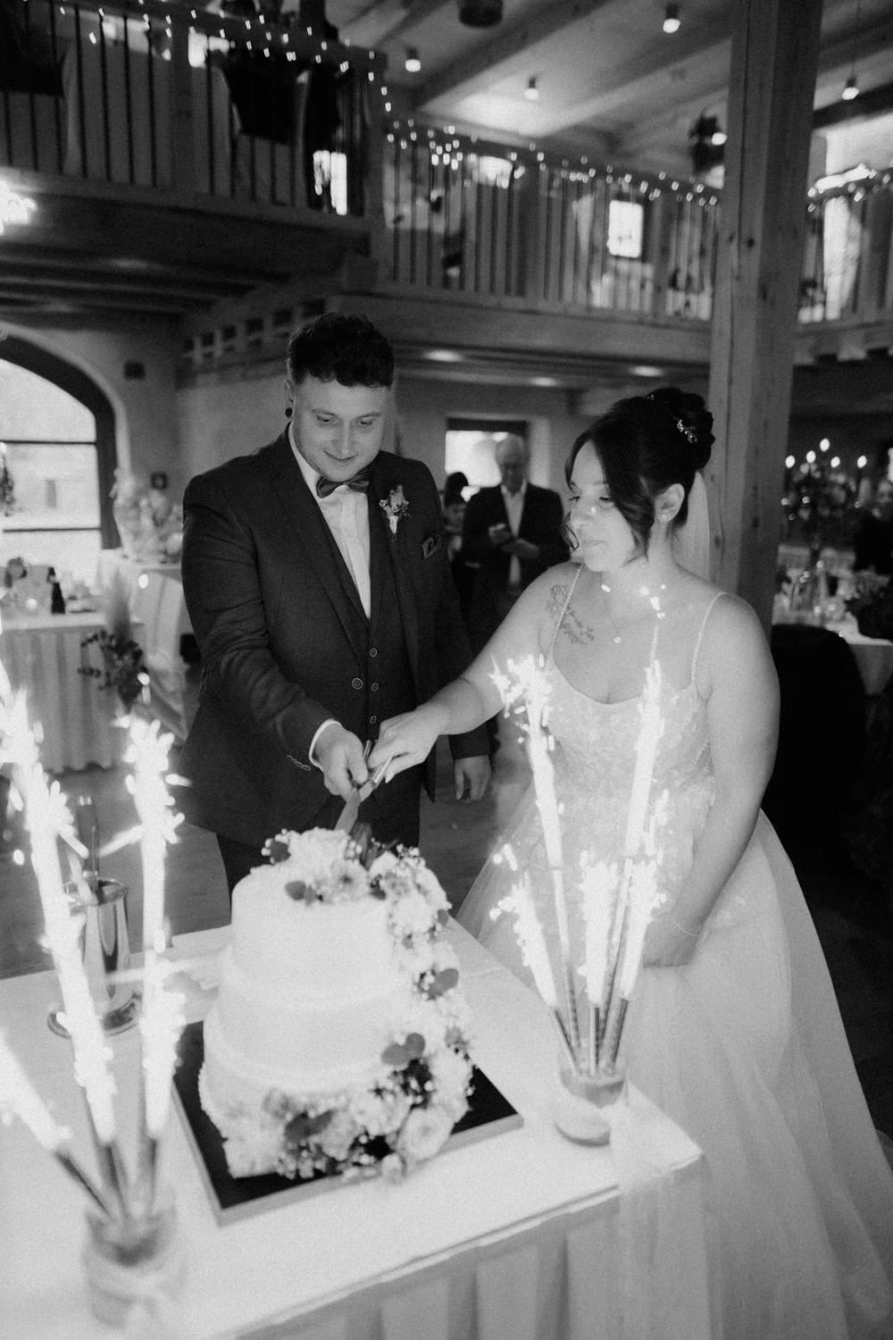 Im Hammerschloss Stodl Schmidmühlen schneidet ein Paar in festlicher Kleidung eine mehrstöckige Hochzeitstorte an, die mit Blumen geschmückt und von Wunderkerzen umgeben ist. Der warm beleuchtete Veranstaltungsort verfügt über elegant dekorierte Tische und ein bezaubernder Balkon darüber trägt zur romantischen Atmosphäre bei.