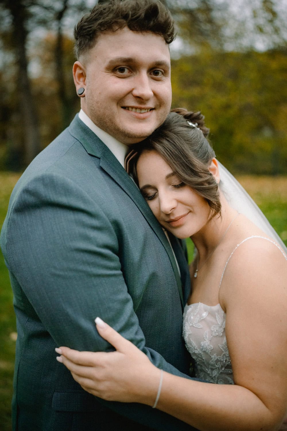Ein Paar umarmt sich liebevoll im Freien vor Hammerschloss Stodl. Der Mann im grauen Anzug lächelt in die Kamera, während die Frau im weißen Kleid ihren Kopf mit geschlossenen Augen auf seiner Brust ruht. Der malerische Hintergrund zeigt grünes Laub und Bäume in Schmidmühlen.