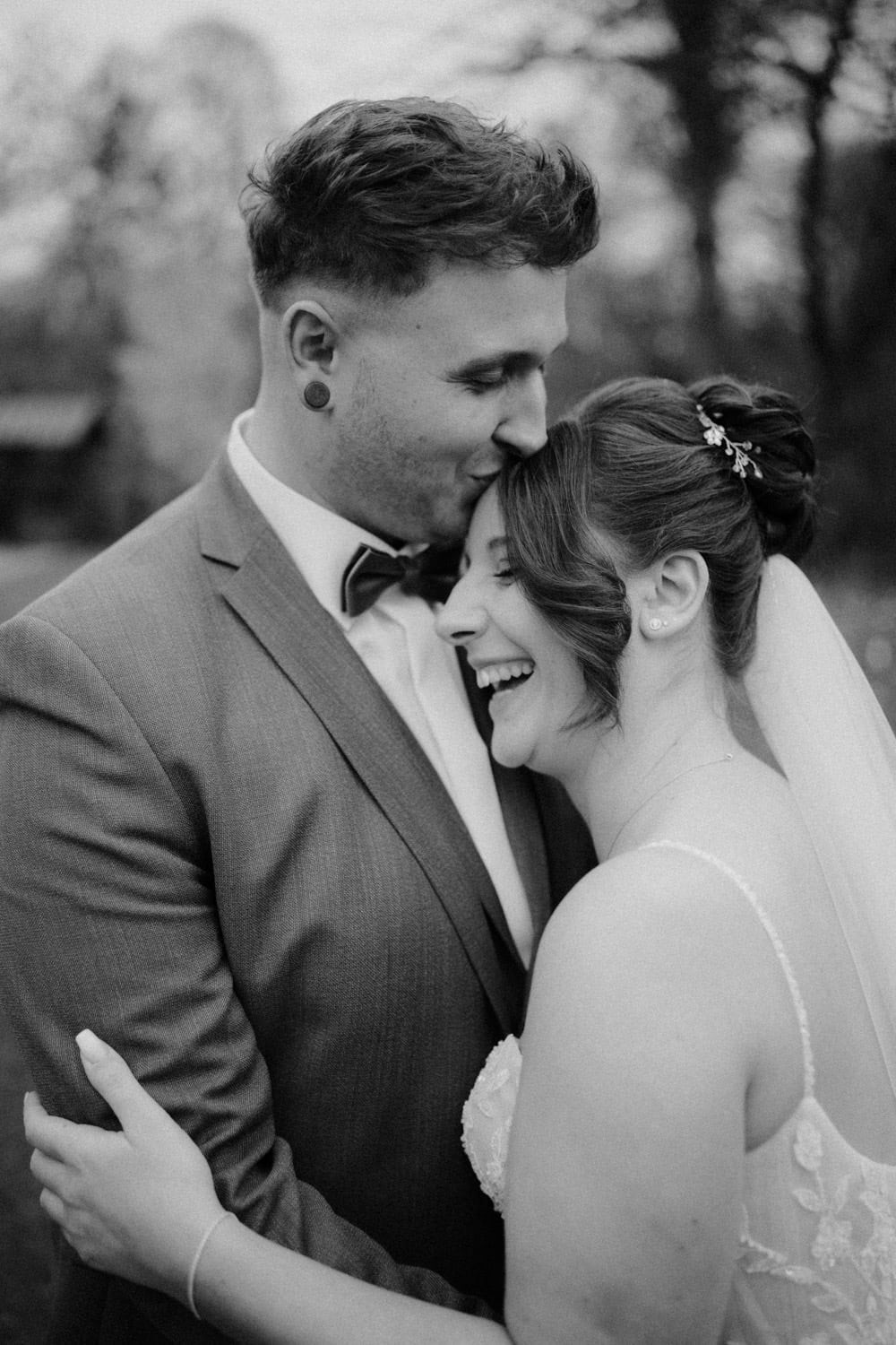 In einem zeitlosen Moment im Hammerschloss Stodl zeigt ein Schwarzweißfoto den Bräutigam im Anzug, wie er seiner Braut im Hochzeitskleid zärtlich die Stirn küsst. Umarmt und lächelnd stehen sie vor einer verschwommenen natürlichen Kulisse.