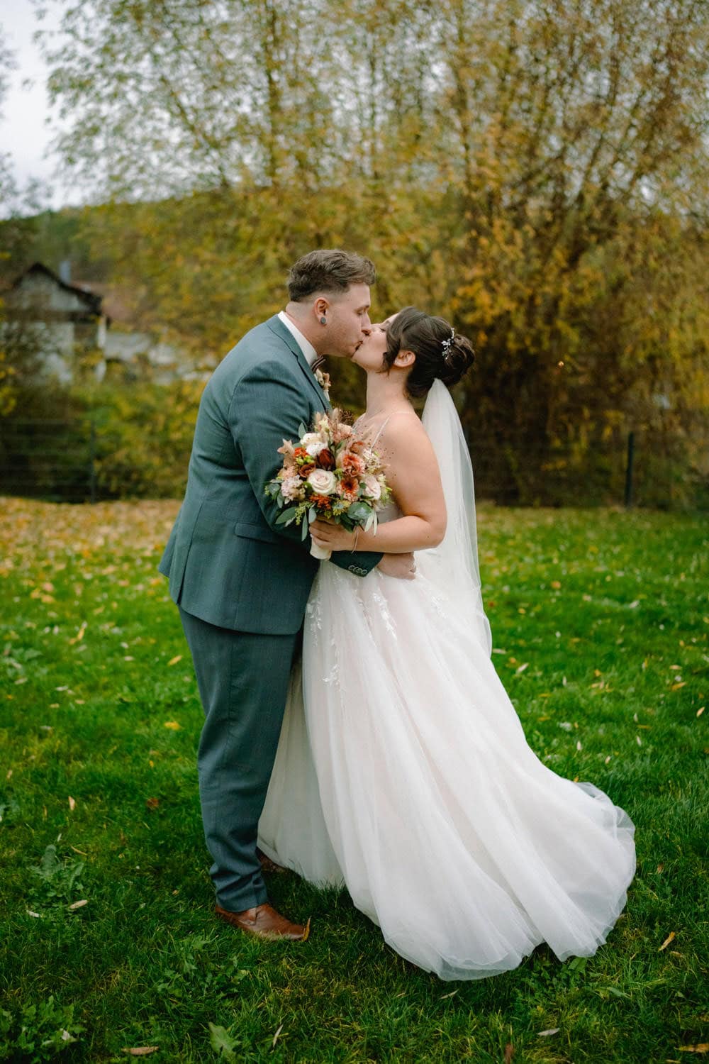 Braut und Bräutigam küssen sich draußen auf der Wiese von Hammerschloss Stodl. Die Braut in ihrem weißen Kleid mit Schleier hält einen Blumenstrauß, während der Bräutigam einen grauen Anzug trägt. Herbstbäume mit gelben Blättern bilden eine malerische Kulisse in Schmidmühlen.