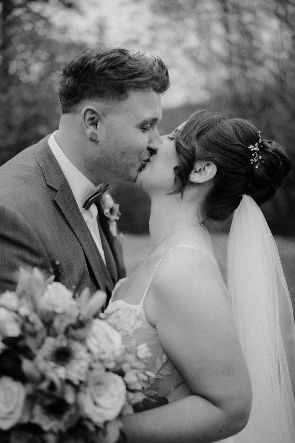 Braut und Bräutigam küssen sich in den bezaubernden Gärten von Hammerschloss Stodl Schmidmühlen. Die Braut hält einen Blumenstrauß und trägt einen Schleier, während der Bräutigam einen Anzug mit Fliege trägt. Bäume bilden den Hintergrund und das Bild ist wunderschön in Schwarzweiß festgehalten.