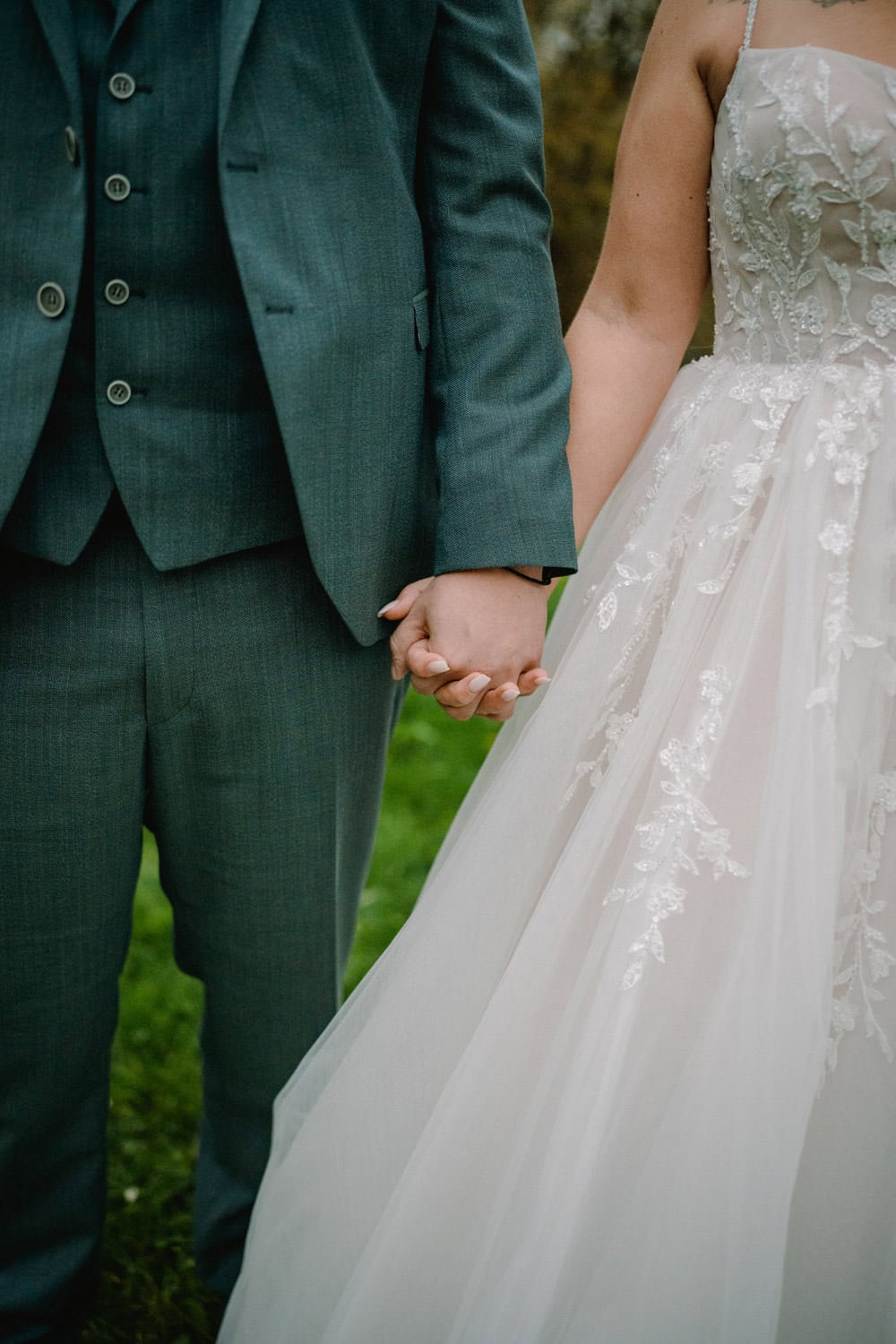 Ein Paar hält sich bei einer Hochzeit im Hammerschloss Stodl Schmidmühlen an den Händen. Der Bräutigam trägt einen grauen Anzug und die Braut ein weißes Spitzenhochzeitskleid. Das Foto konzentriert sich auf ihre gefalteten Hände und hebt die Details ihrer Kleidung vor dem grasbewachsenen Hintergrund hervor.