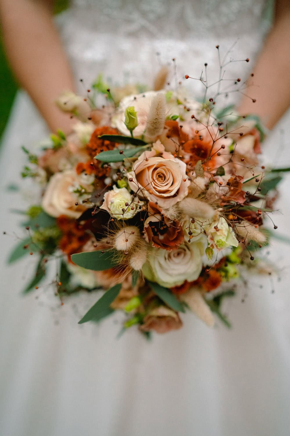Eine Person in einem weißen Kleid hält einen Strauß Rosen und Grünpflanzen im Hammerschloss Stodl Schmidmühlen. Das Blumenarrangement mit sanften, gedämpften Tönen und Ziergräsern strahlt Eleganz und natürliche Schönheit inmitten des historischen Charmes des Ortes aus.