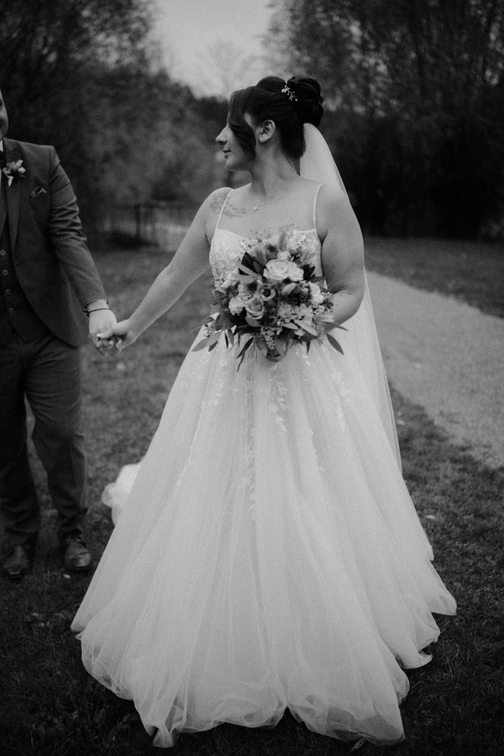 Eine Braut im wallenden Brautkleid hält einen Blumenstrauß und die Hand ihres Partners. Sie stehen auf einem Grasweg, umgeben von Bäumen beim Hammerschloss Stodl Schmidmühlen. Die bezaubernde Szene ist in zeitlosem Schwarzweiß festgehalten.