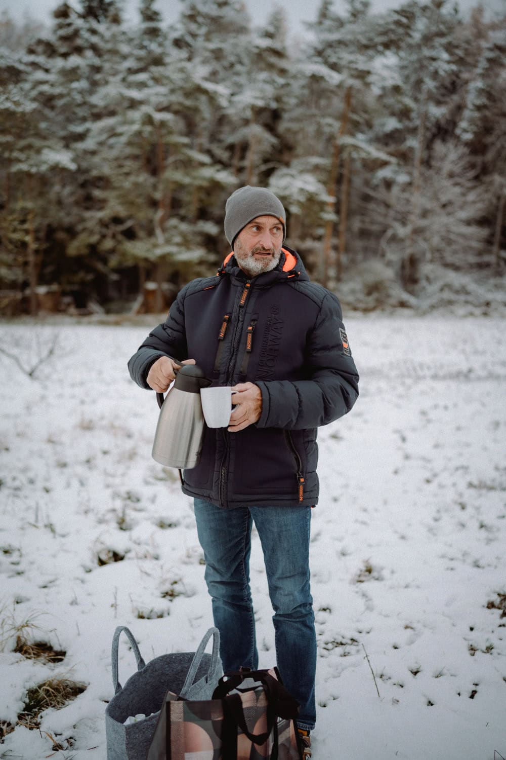 Ein Mann steht auf einem schneebedeckten Feld in der Nähe von Hammerschloss Stodl. Er trägt eine graue Mütze und eine schwarze Winterjacke. Er gießt ein heißes Getränk aus einer Thermoskanne in eine Tasse. In der Nähe liegen Taschen im Schnee, während Waldbäume die Szene einrahmen und im Hintergrund der Charme von Schmidmühlen zu spüren ist.