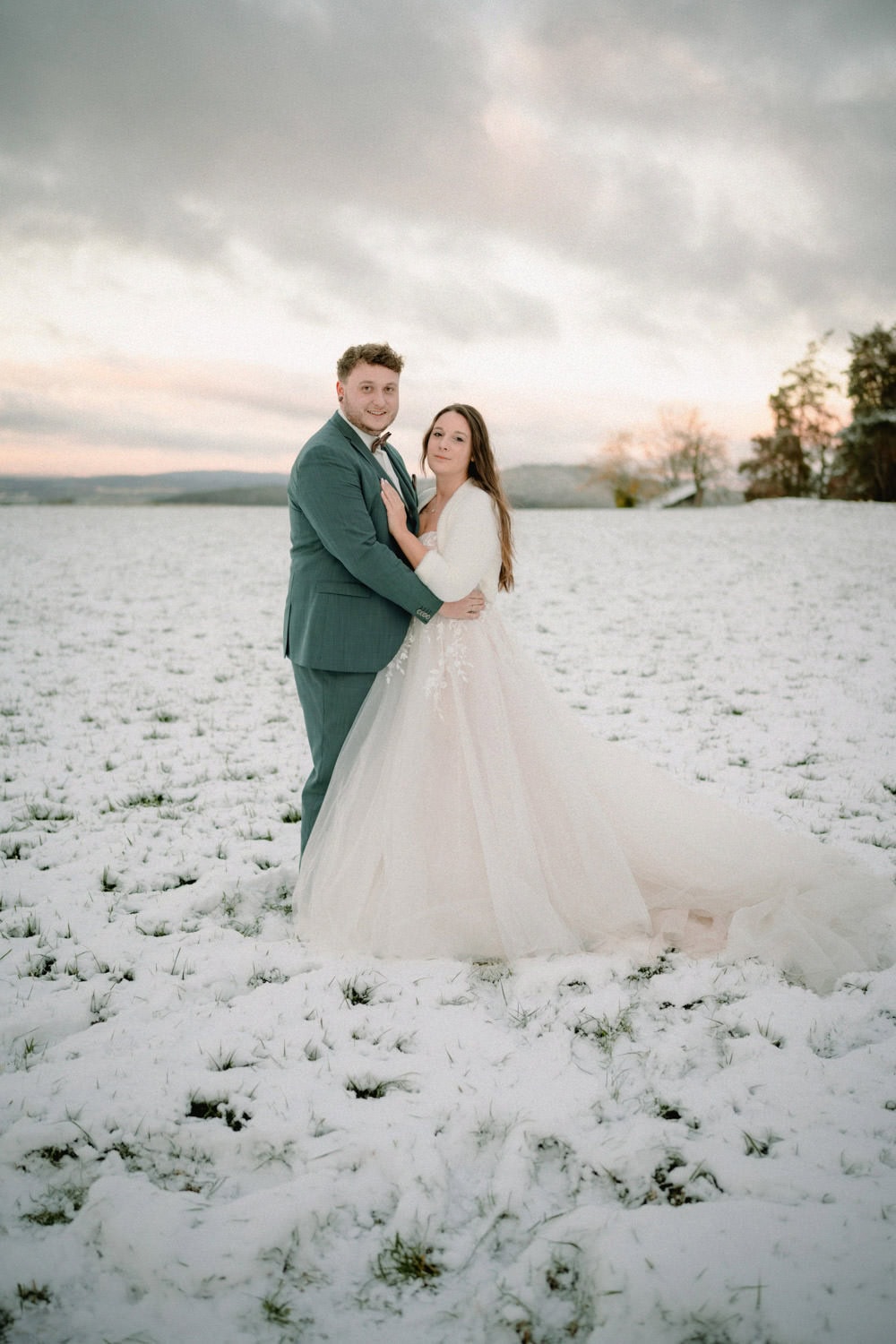 Ein Paar steht auf einem schneebedeckten Feld in der Nähe des bezaubernden Hammerschlosses Stodl Schmidmühlen und umarmt sich. Der Bräutigam trägt einen grünen Anzug und die Braut ein fließendes weißes Kleid. Der bedeckte Himmel hat bei Sonnenuntergang einen Hauch von Rosa und Orange und wirft einen romantischen Glanz in den Hintergrund.