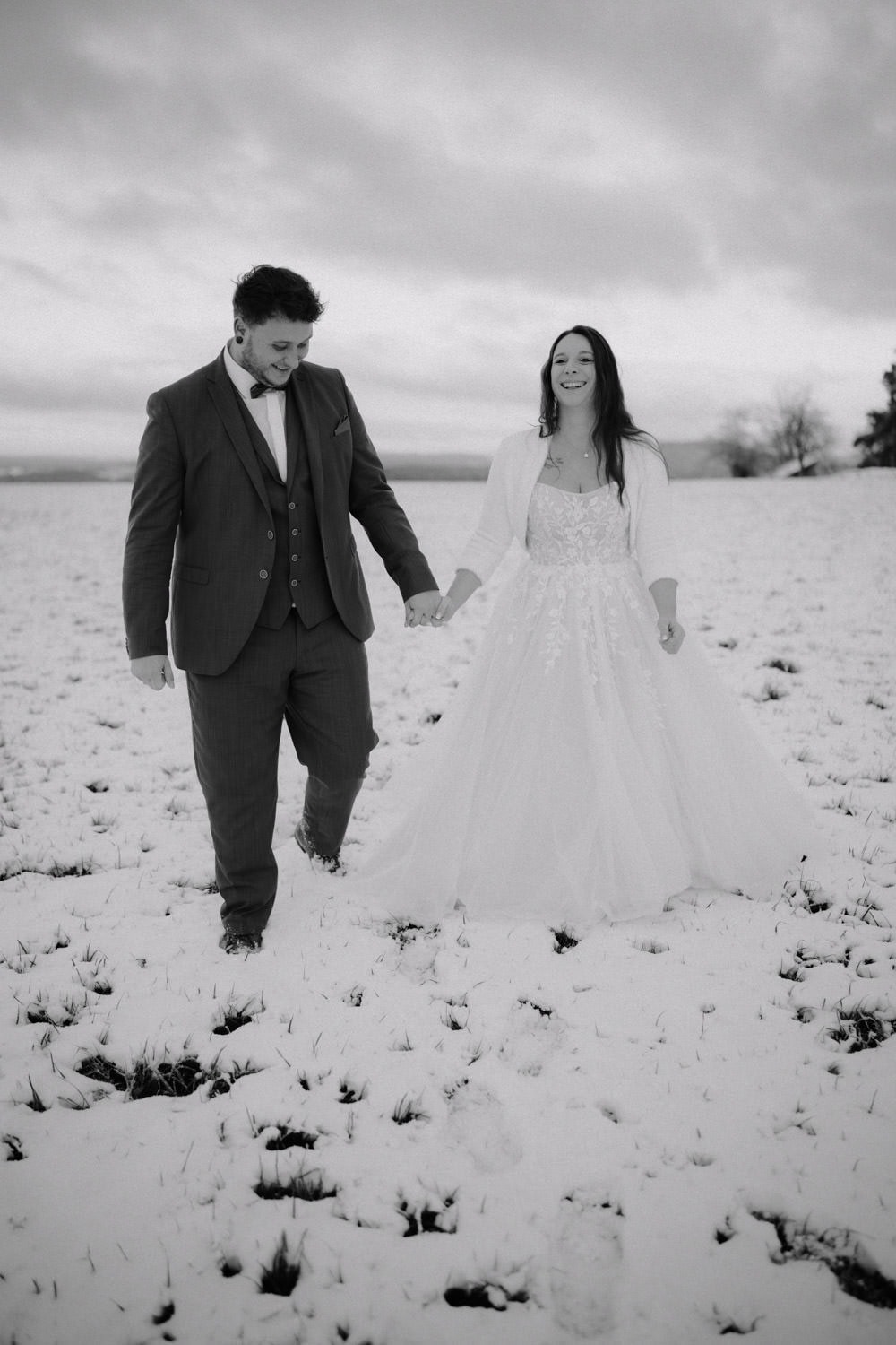 Ein Paar in Abendgarderobe, der Bräutigam im Anzug und die Braut im weißen Brautkleid, gehen Hand in Hand durch ein schneebedecktes Feld in der Nähe von Hammerschloss Stodl. Der Himmel ist bewölkt und in der Ferne rahmen Bäume die heitere, freudige Szene ein.