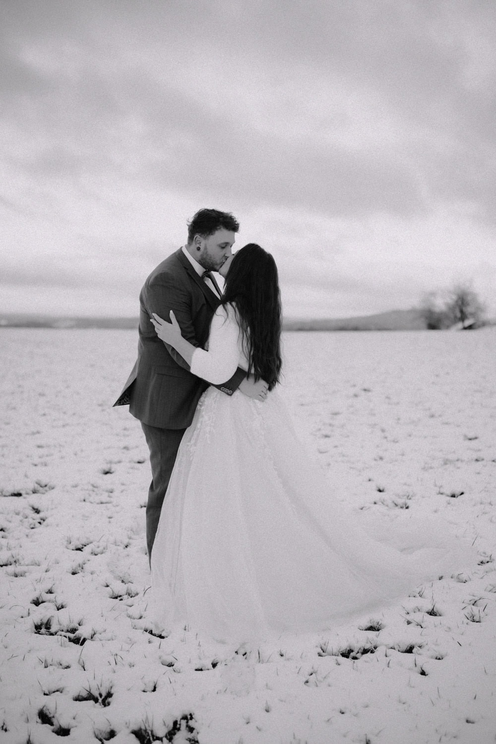 Ein Paar umarmt sich auf einem schneebedeckten Feld in der Nähe von Hammerschloss. Die Braut trägt ein langes Kleid und der Bräutigam einen Anzug. Die Landschaft in der Nähe von Stodl Schmidmühlen ist schneebedeckt, während ein wolkenverhangener Himmel darüber schwebt.