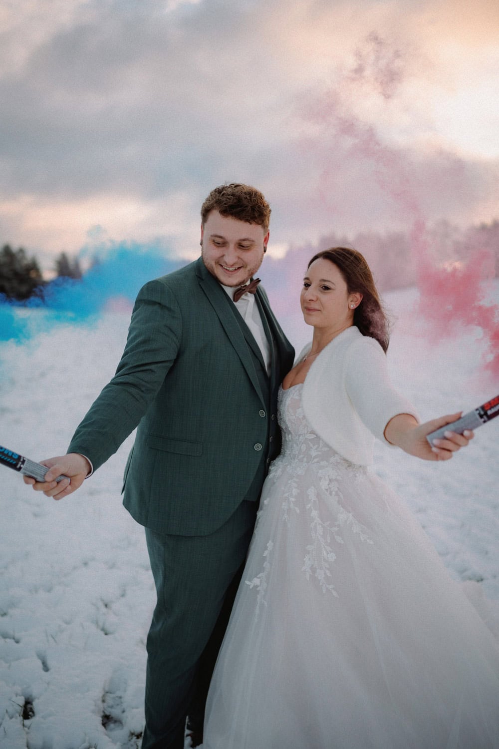 Braut und Bräutigam stehen gemeinsam in einer verschneiten Landschaft beim Hammerschloss Stodl Schmidmühlen. Beide halten jeweils einen Rauchstab in der Hand, aus dem bunter rosa und blauer Rauch aufsteigt. Die Braut trägt ein weißes Kleid mit einem Schal und der Bräutigam einen dunklen Anzug. Beide lächeln, während im Hintergrund die Sonne untergeht.