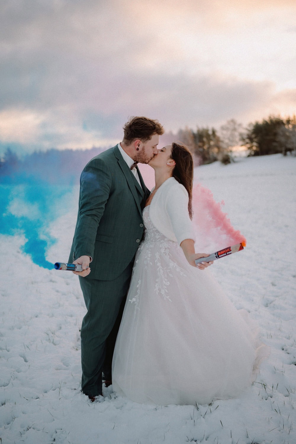 Auf dem verschneiten Feld von Hammerschloss Stodl Schmidmühlen küsst sich ein Paar in Hochzeitskleid und Anzug leidenschaftlich und hält Rauchbomben in den Händen, aus denen blauer und rosa Nebel strömt. Hinter ihnen rahmen Bäume die Szene vor einem wolkigen Himmel ein, der in den warmen Farben des Sonnenuntergangs gefärbt ist.
