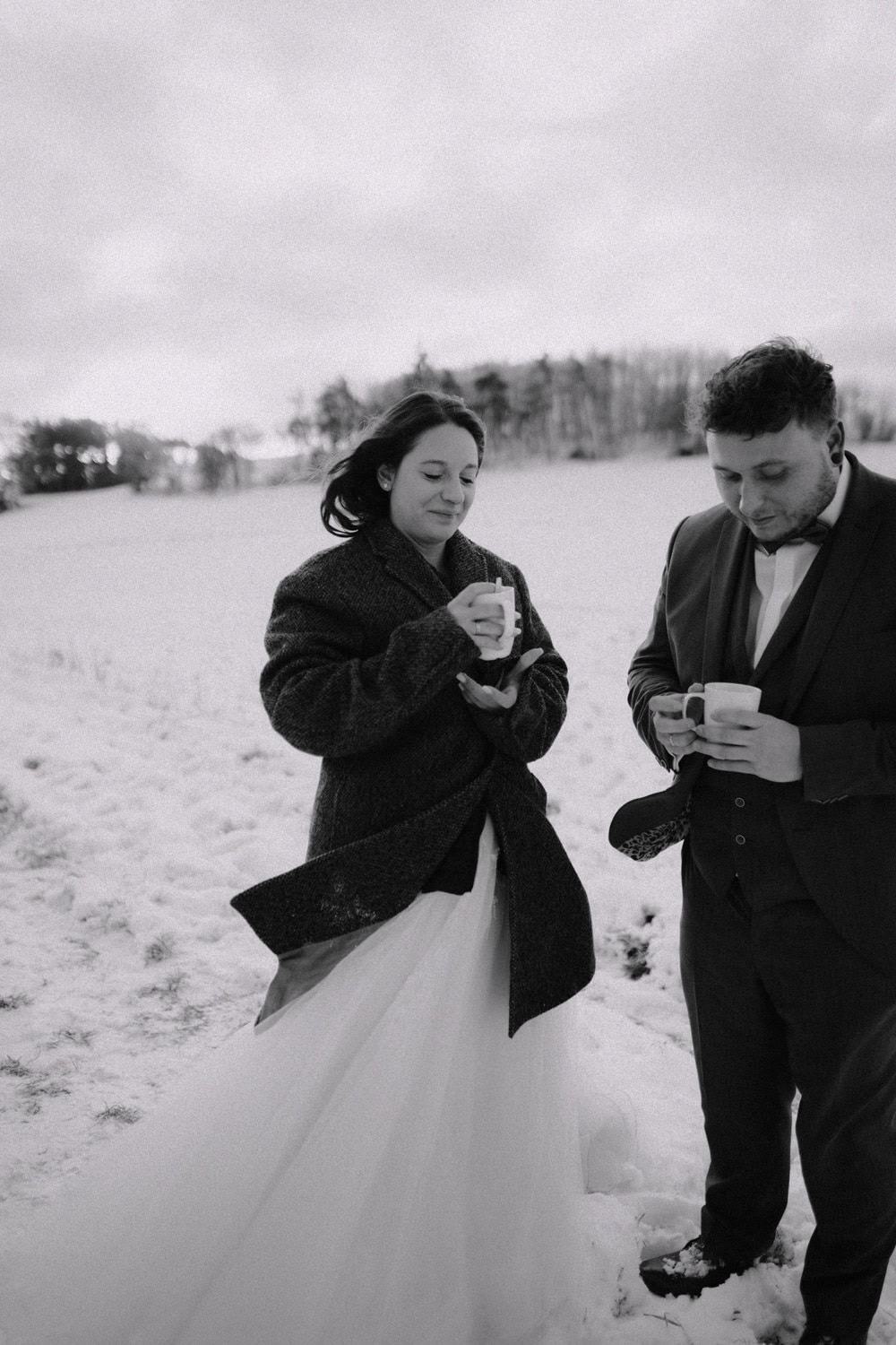 Auf einem zeitlosen Schwarzweißfoto stehen Braut und Bräutigam draußen vor Hammerschloss Stodl Schmidmühlen, beide halten einen Becher in der Hand, auf dem schneebedeckten Feld. Die Braut trägt ein Abendkleid unter ihrem dunklen Mantel, der Bräutigam ist elegant in seinem Anzug. Hinter ihnen ragen kahle Bäume in den wolkigen Himmel.