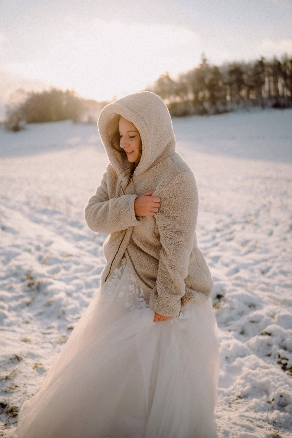 Eine Person in einem weißen Gewand und einer kuscheligen Kapuzenjacke steht auf einem schneebedeckten Feld in der Nähe von Hammerschloss, Schmidmühlen. Die Sonne geht dahinter unter und wirft einen warmen Schein über die Winterlandschaft. Im Hintergrund sind Bäume zu sehen, die die ruhige Szenerie noch verstärken.