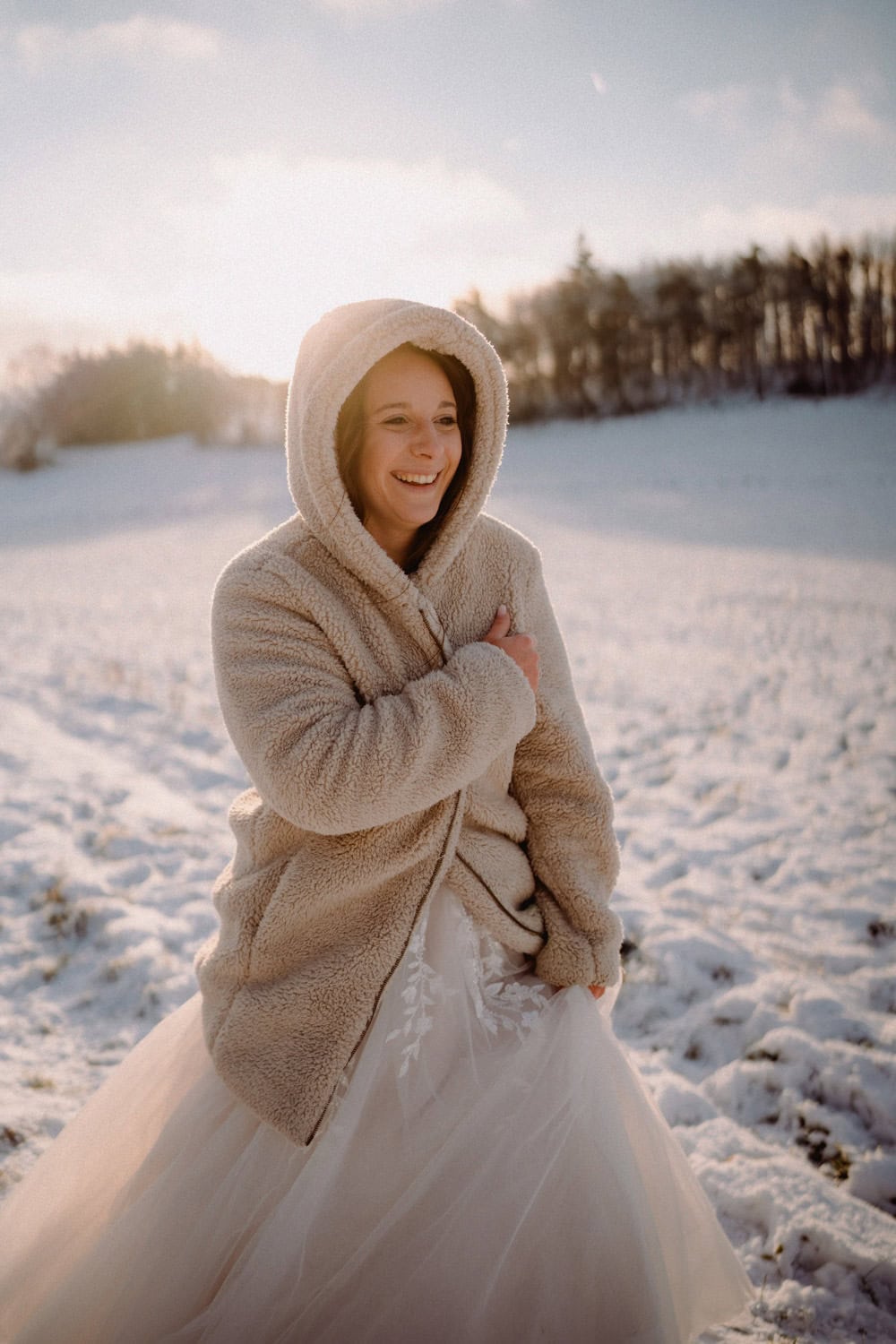 Eine Frau in einem Brautkleid und einer kuscheligen Kapuzenjacke lächelt, während sie in den schneebedeckten Feldern von Hammerschloss Stodl Schmidmühlen steht, hinter ihr geht die Sonne unter und in der Ferne sind Bäume zu sehen.