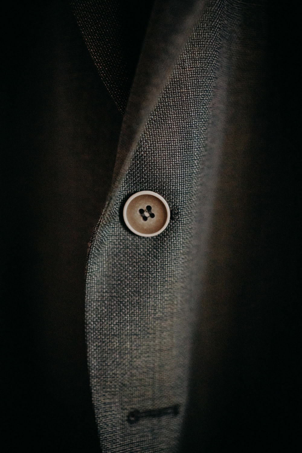 Nahaufnahme eines einzelnen braunen Knopfs auf einem strukturierten grauen Stoff aus der klassischen Kollektion von Hammerschloss Stodl Schmidmühlen, möglicherweise Teil eines Anzugs oder Mantels. Der Stoff ist fein gewebt und der Knopf ist durch ein Knopfloch befestigt, was zeitlose Eleganz ausstrahlt.