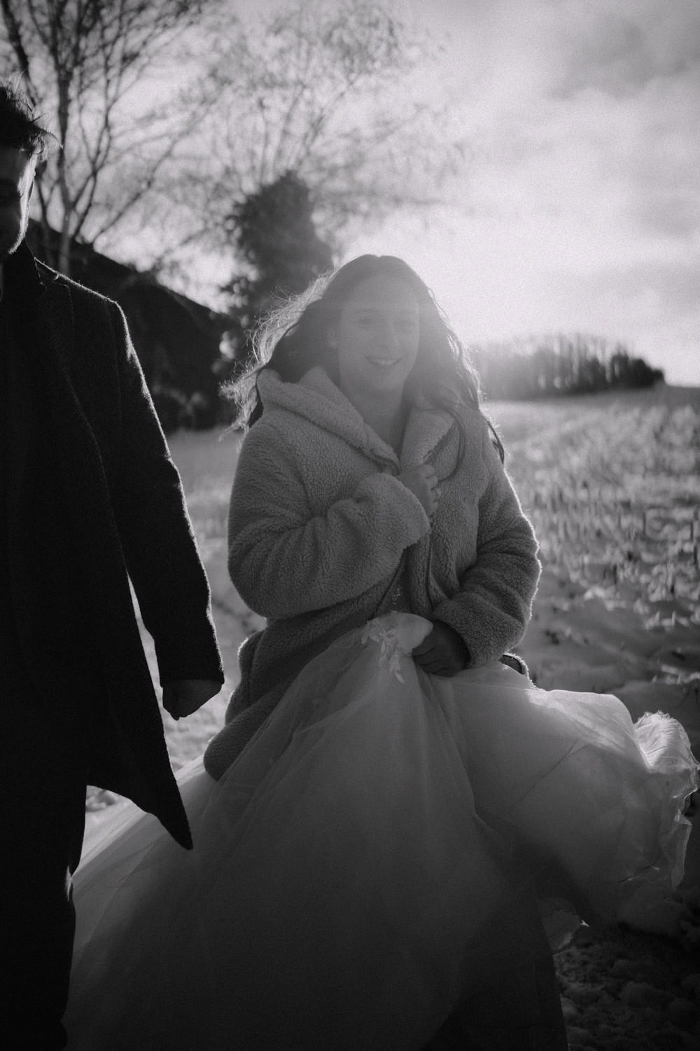 Eine Frau in einem flauschigen Mantel und einem wallenden Kleid geht mit einer Person an ihrer Seite durch ein schneebedecktes Feld in der Nähe von Hammerschloss Stodl. Die Sonne geht hinter Schmidmühlen unter und wirft warmes Licht und lange Schatten über die bezaubernde Szene.