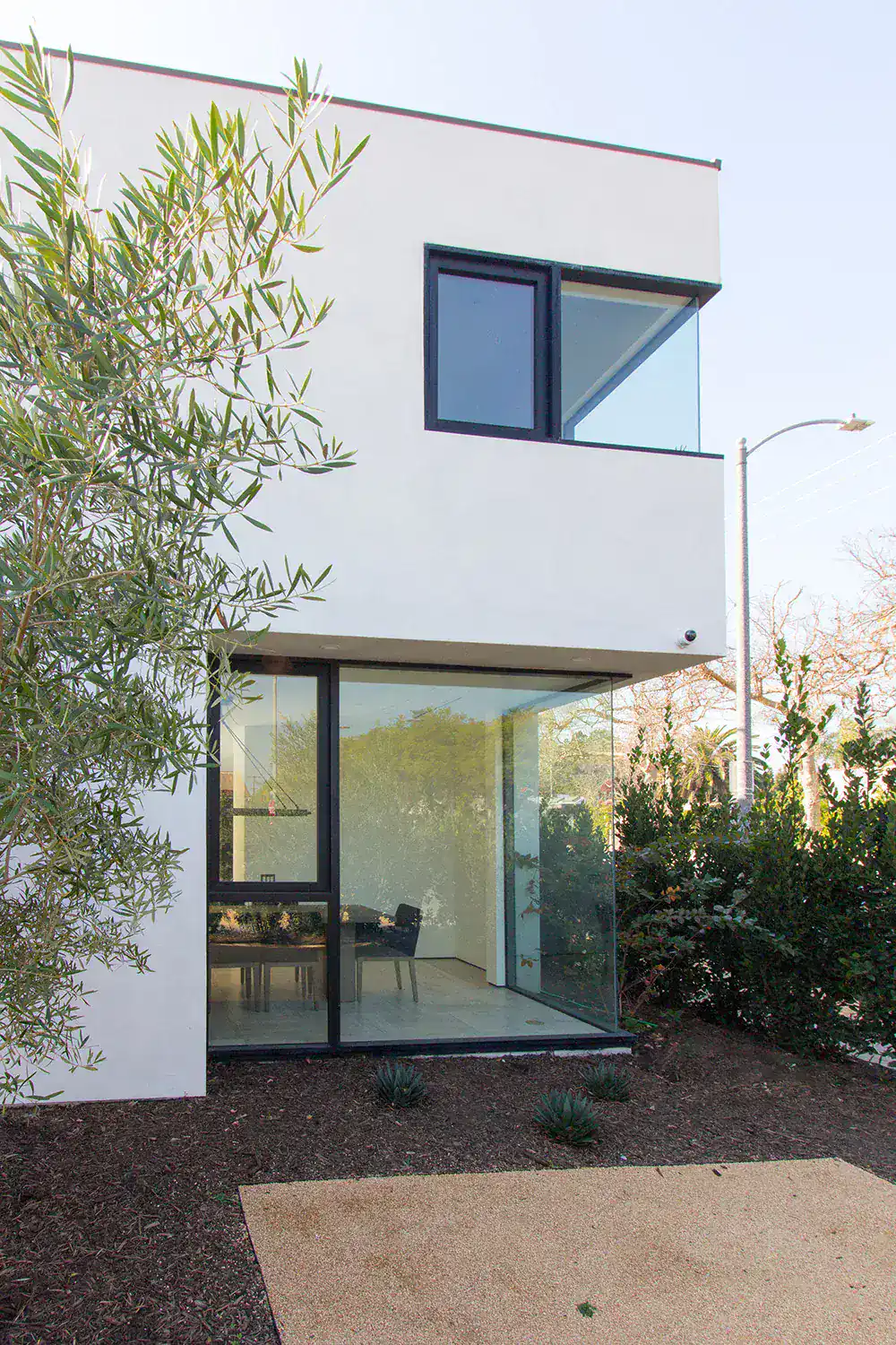 Modern minimalist house with white facade, large glass windows, and sleek black trim, surrounded by greenery and natural landscaping.