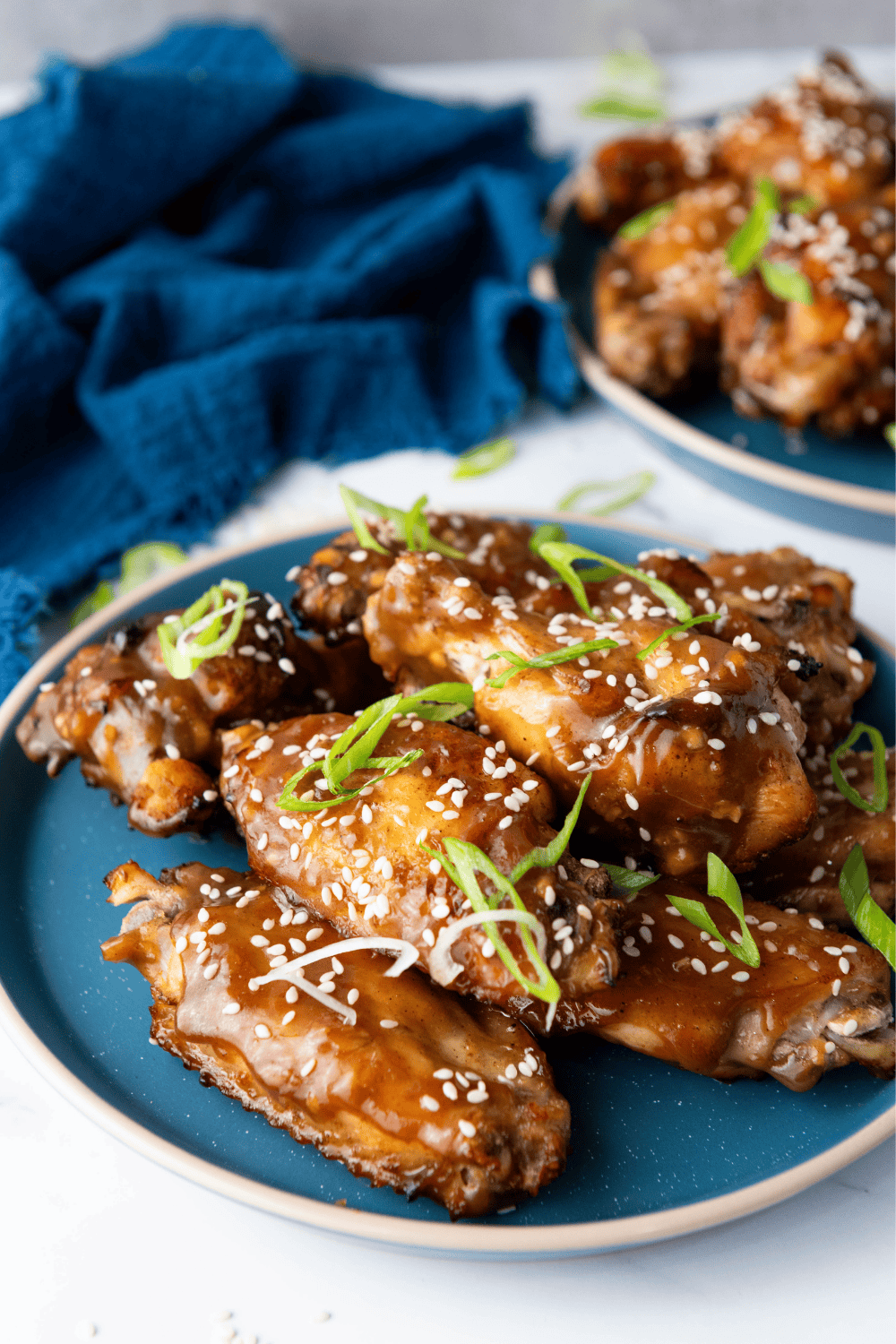A close-up view of sticky Chinese Chicken Wings garnished with sesame seeds and green onions on a blue plate, set against a dark blue cloth background.