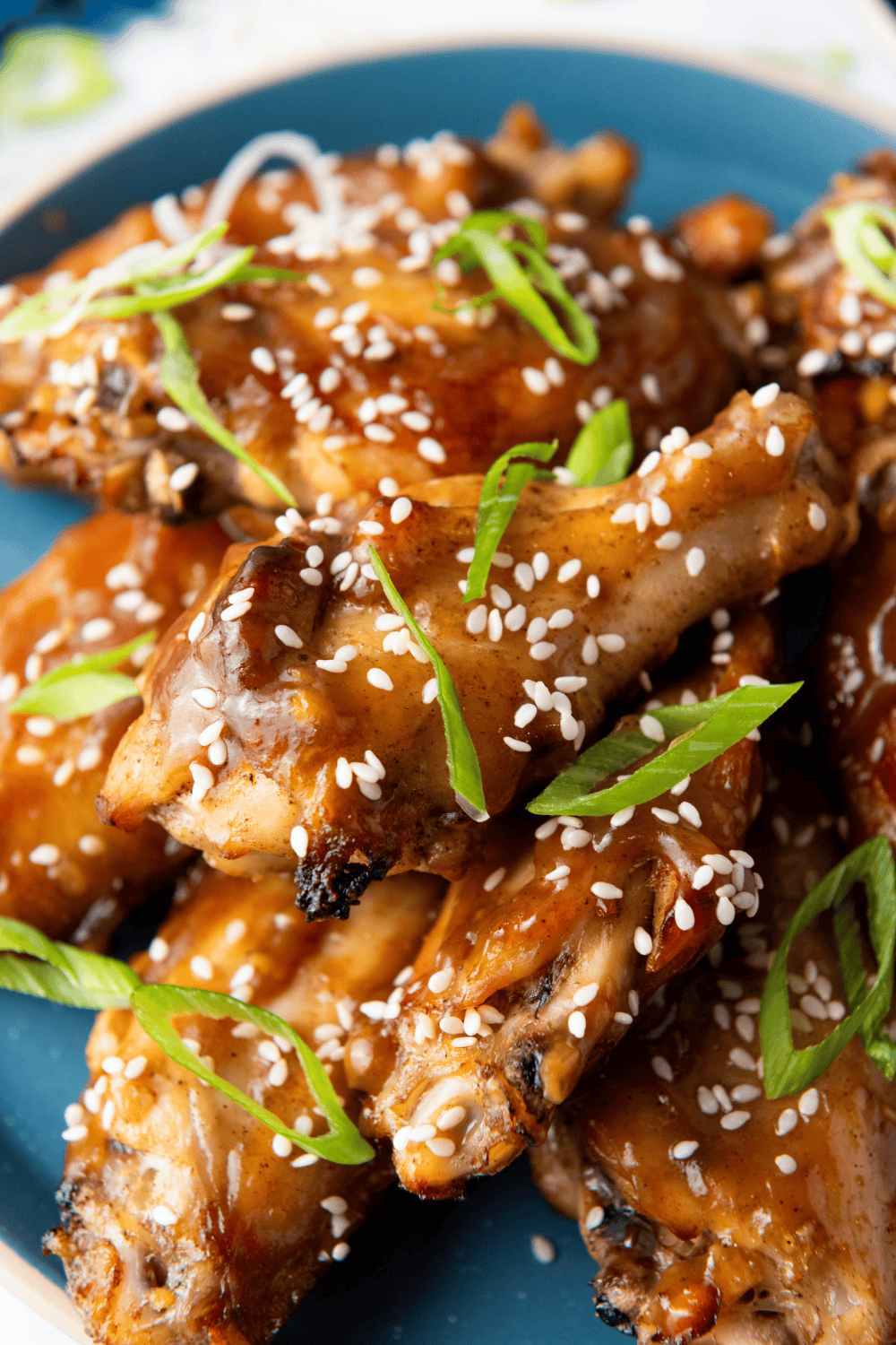 A vertical extreme close-up focusing on the glossy, sticky glaze of two Chinese Chicken Wings, revealing the crispy skin and numerous sesame seed and green onion toppings.