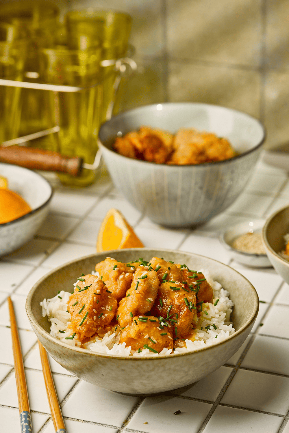 A bowl of orange chicken on rice in the foreground with a blurry bowl and kitchen items in the background.