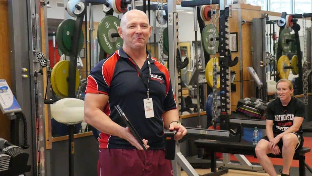 A strength and conditioning coach standing confidently in a gym, clipboard in hand.