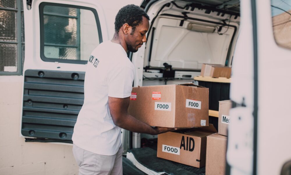 man placing food labelled carboard boxes inside the trunk of a white van