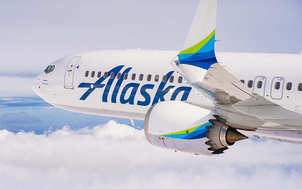 Alaska Airlines airplane flying above clouds with the word “Alaska” visible on the side and blue and green accents on the wing and engine.