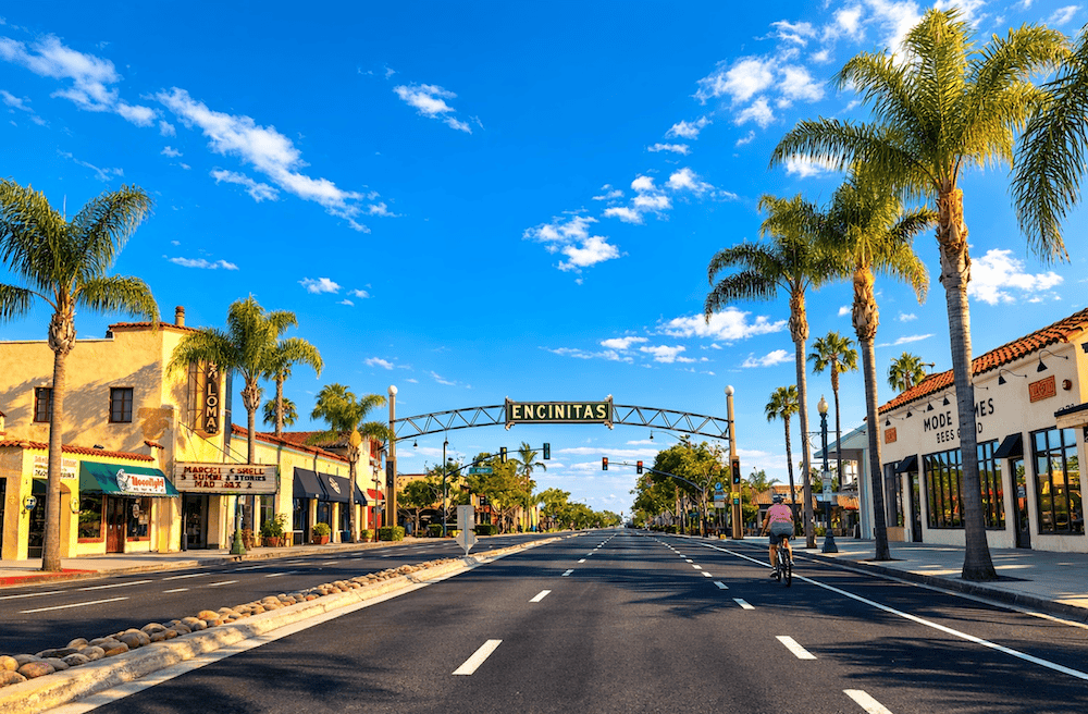 Car-free view of downtown Encinitas with the iconic Encinitas arch, palm trees, Spanish-style storefronts, bright blue sky, and sunny coastal California atmosphere.