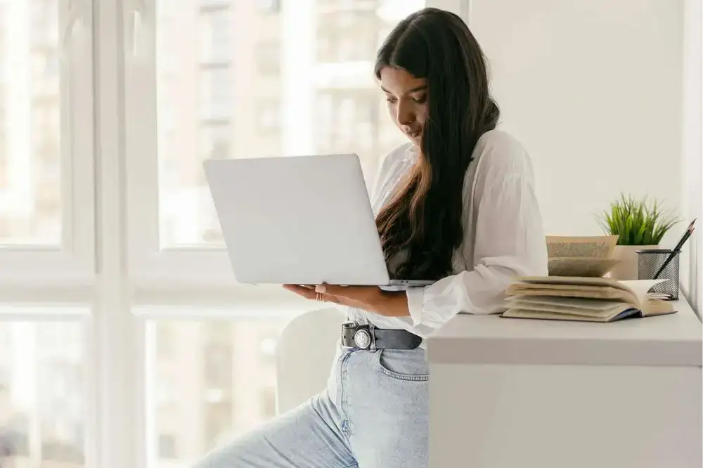 Woman leaning on a white desk while using a silver laptop, with books and a plant nearby in a bright room representing the benefits of EMDR therapy in Michigan and the healing journey through EMDR in Ann Arbor, MI.