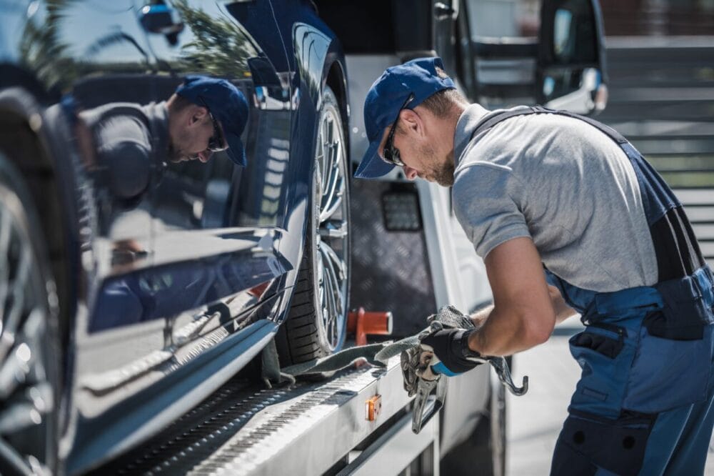 towing edmonton A Towing Edmonton FLM tow truck operator in uniform secures a blue car onto a flatbed using straps, working outdoors on a sunny day. towing edmonton