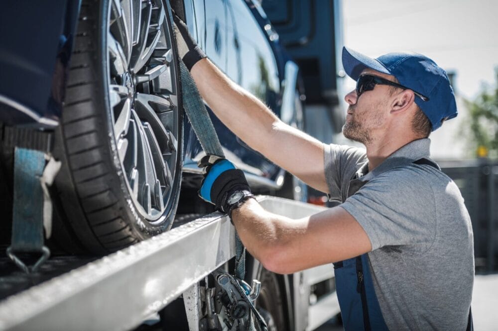towing edmonton A man wearing a blue cap, sunglasses, and gloves secures a car wheel with straps onto a vehicle transport trailer on a sunny day, showcasing professional towing Edmonton FLM service. towing edmonton