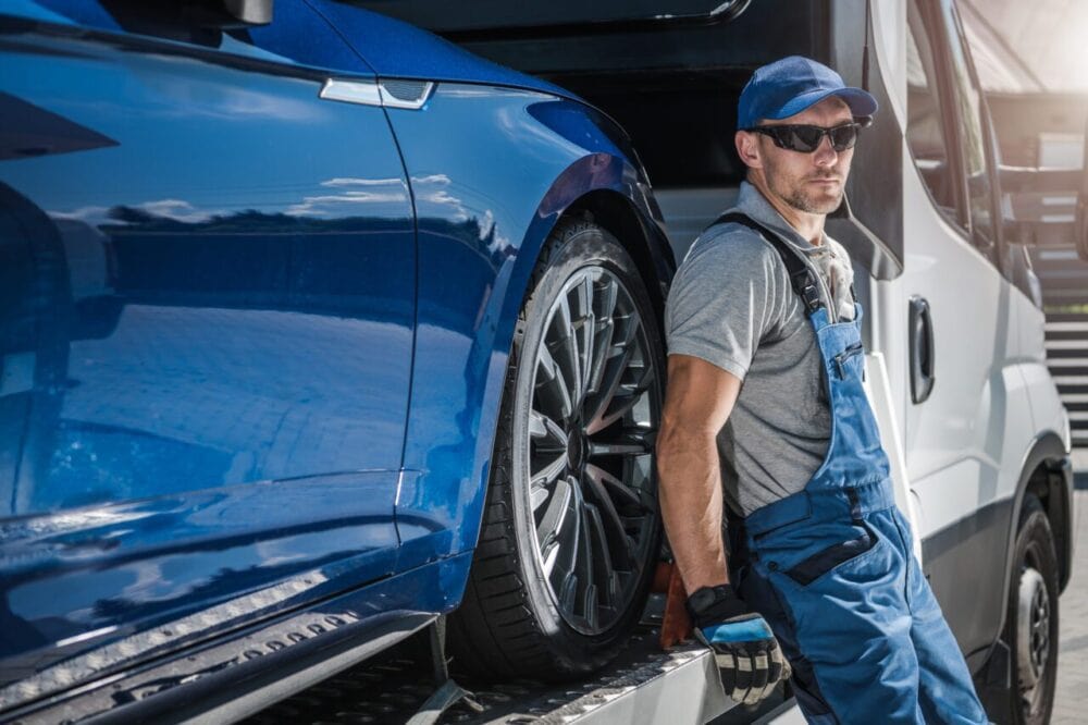 towing edmonton A mechanic in blue overalls, cap, and sunglasses leans against a blue car loaded on a Towing Edmonton FLM truck, looking at the camera with a serious expression. The scene appears to be outdoors on a sunny day. towing edmonton