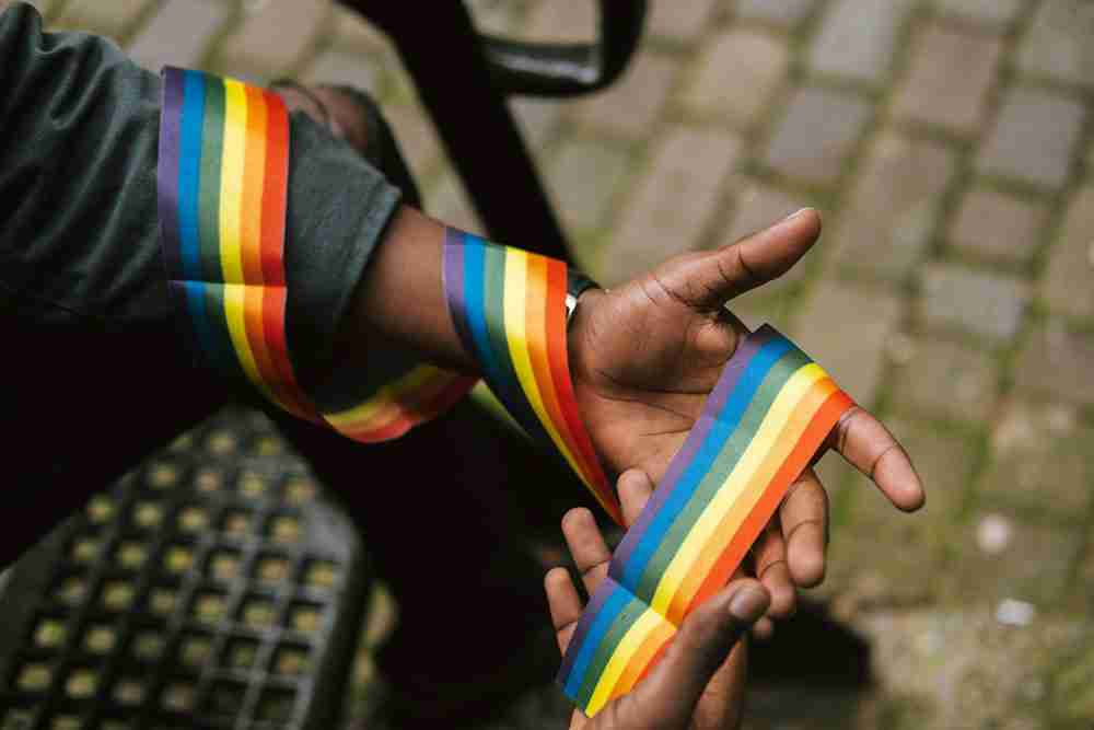 Two hands wrap a rainbow Pride ribbon around their wrists, celebrating LGBTQ+ pride in a close-up shot on a cobblestone surface. See an LGBTQ Therapist in Detroit today!