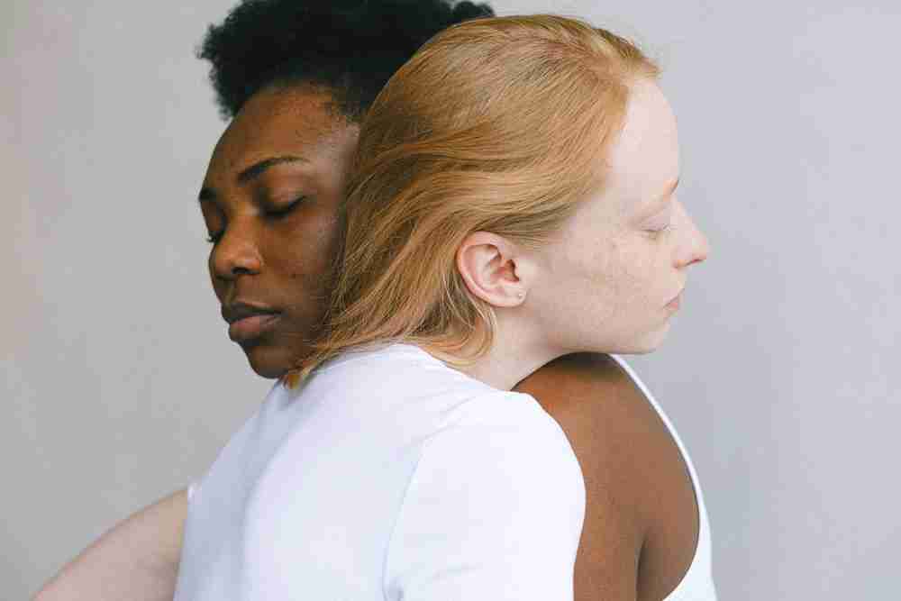 Two women hug back-to-back with eyes closed; one has dark skin and short hair, the other is light-skinned with blonde hair in white tops symbolizing empowerment and healing through LGBT therapy and LGBT trauma therapy in Ann Arbor, MI.