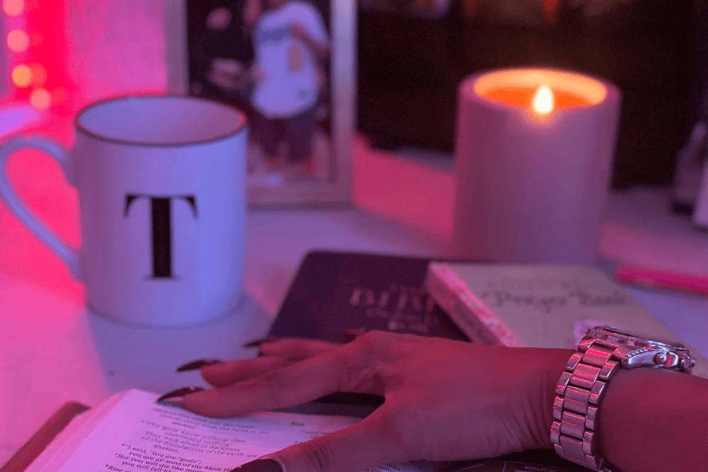 A serene devotional setup featuring an open Bible highlighted in pink lighting, with a hand resting gently on its pages. A lit candle, a "Prayer Journal," a mug with the letter "T," and a framed photograph in the background create a peaceful atmosphere. The image symbolizes faith and trust in manifesting what’s already destined for you.
