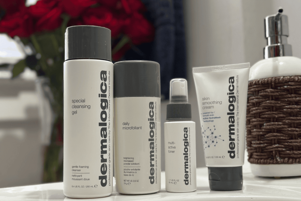A neatly arranged collection of Dermalogica skincare products on a clean white surface, with a background featuring vibrant red roses and soft lighting. The products include a special cleansing gel, skin moisturizer, smoothing cream, and a mist spray, symbolizing self-care and winter skincare essentials.