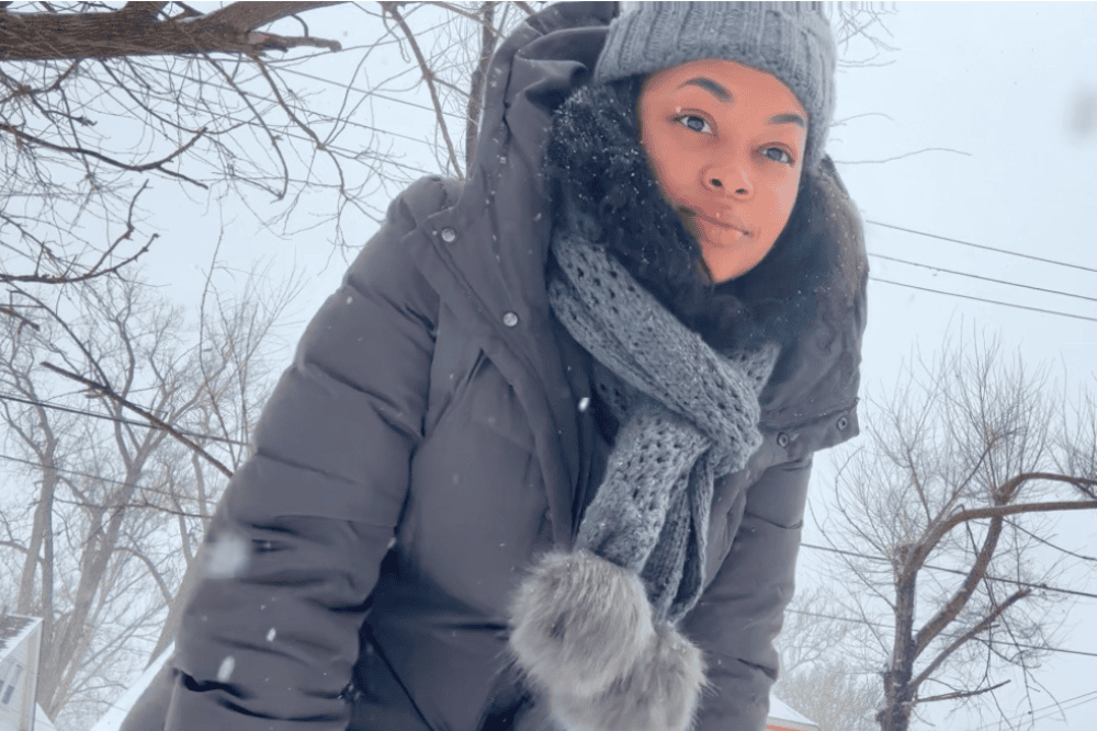 A woman standing outside during a gentle snowfall, wearing a gray coat, hat, and scarf. She exudes warmth and comfort in the cold, embodying self-care and wellness during the winter season. Snowflakes fall softly, adding a serene and refreshing winter backdrop.