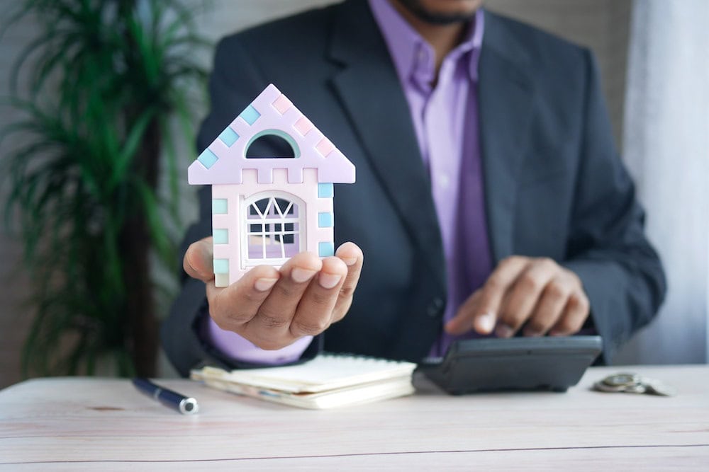 A man holding a small mockup of a house while using a calculator on the desk
