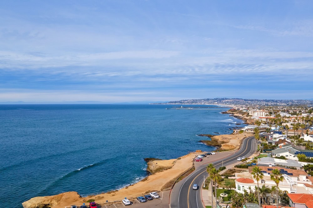 Aerial view of the Ocean Beach coastline in San Diego, featuring a winding coastal road, rocky cliffs, and the Pacific Ocean under a blue sky with light clouds.