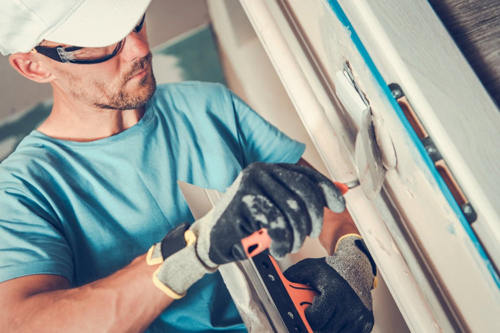 A professional handyman in Coeur D'Alene, Idaho, wearing safety glasses and gloves, uses a putty knife to apply filler to a wall for home repairs.