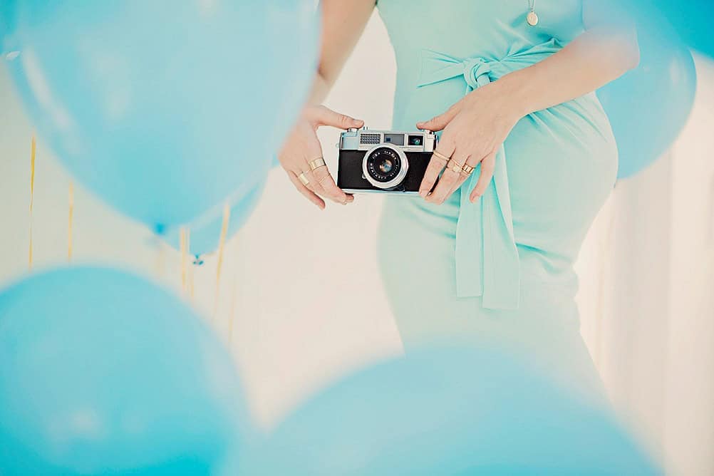 Nydelig gravidfotografering med blå ballonger og gravid kvinne i lys blå kjole, som holder et vintage kamera, perfekte bilder for rammetema gravidfoto.