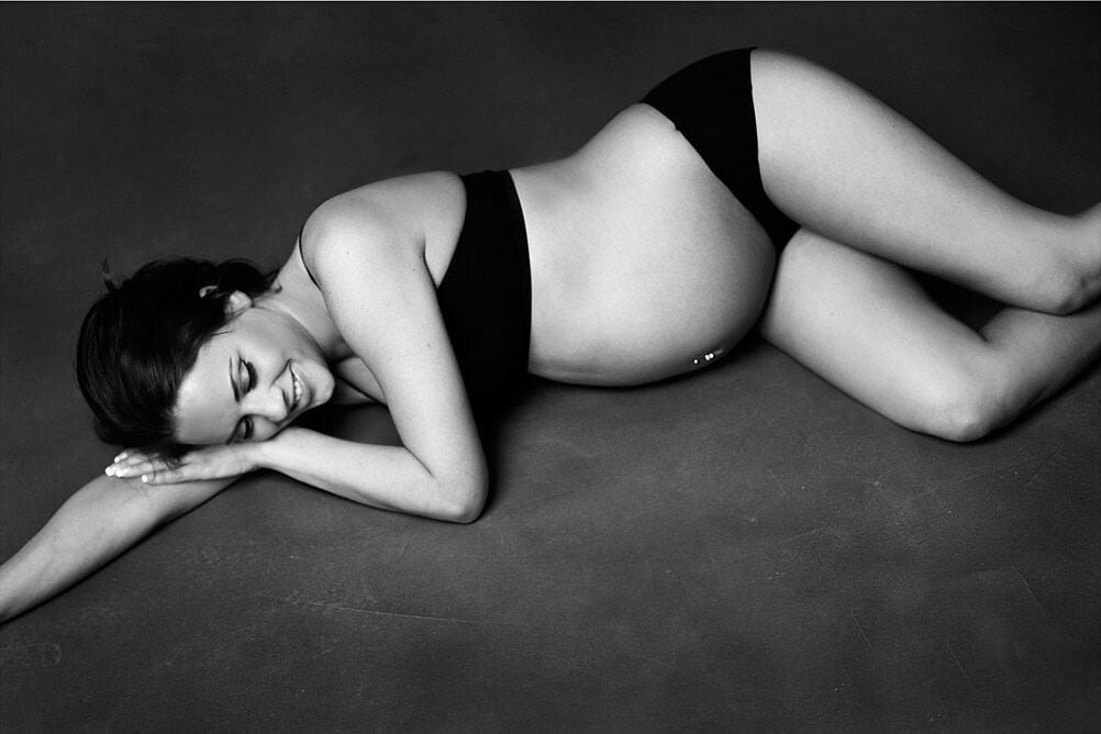 Nydelig gravide kvinne i svart undertøy, liggende på magen med et smilende ansikt, i en nyfødt gravidfoto-stil for å feire mor- og farskapet.