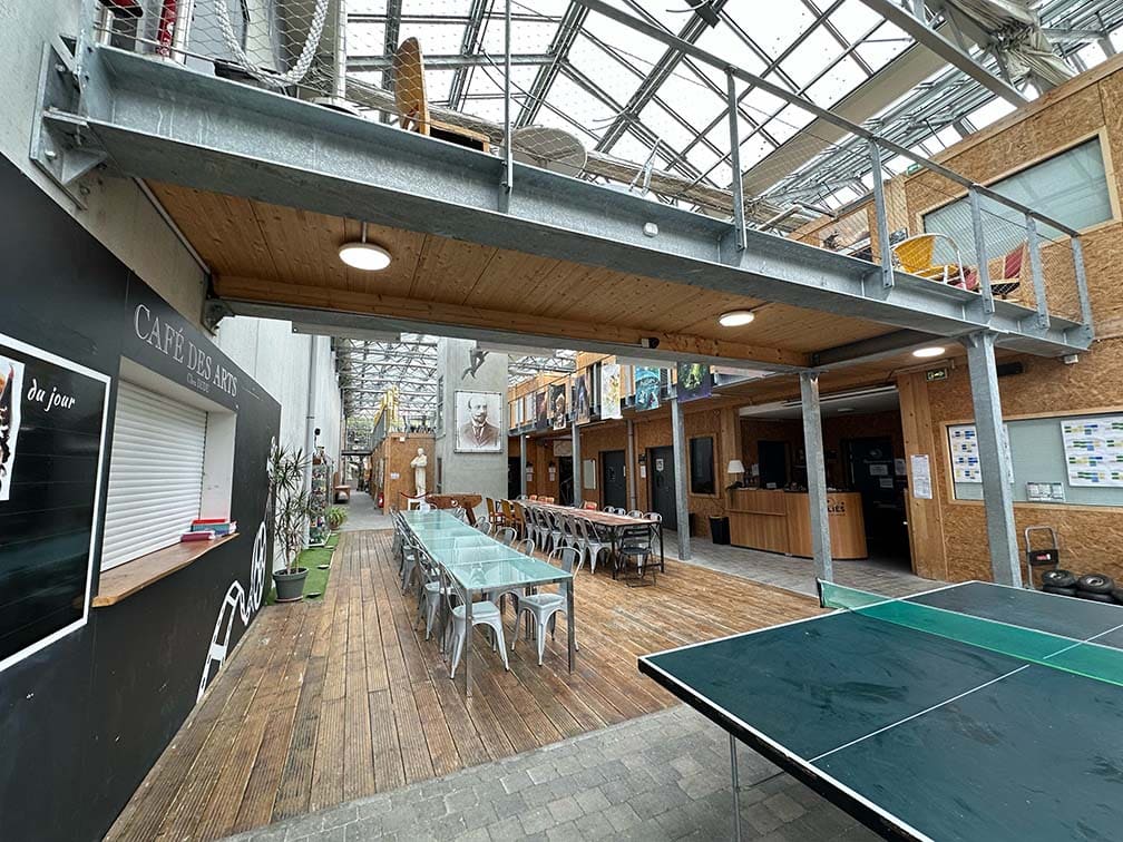 L’atrium de l’École Méliès sous sa verrière : de longues tables et des chaises sur un plancher de bois, le comptoir du Café des Arts à gauche, une table de ping-pong au premier plan et une passerelle en mezzanine