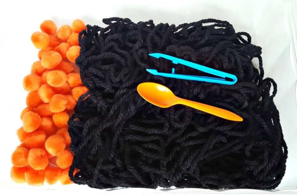 Halloween themed sensory bin featuring black yarn and orange pom poms