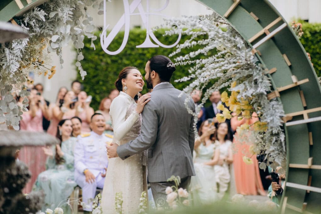 Nagaya and Amarvir share a laugh in front of their guests under a floral arch at their Thai wedding at U Sathorn Bangkok