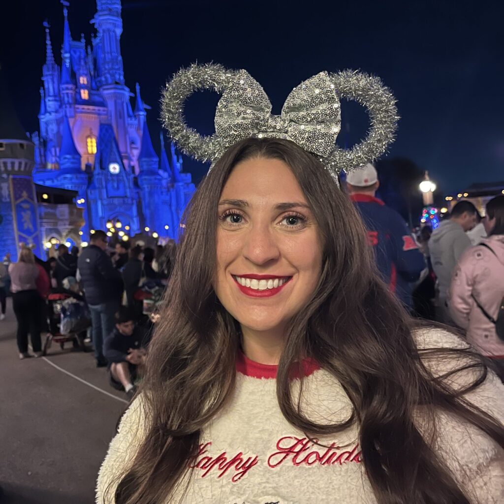 Woman smiling at Disney park with castle in background, wearing holiday sweater and Mickey ears.