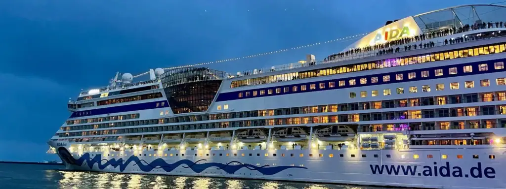 csm_AIDAdiva_Warnemuende__3__56f93ead14 - Michis Kreuzfahrten das kreuzfahrtschiff aidadiva vor dem hafen bei abenddämmerung, beleuchtet mit bunten lichtern und reflektiert im wasser.