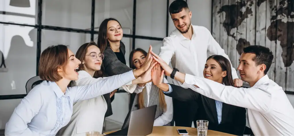 A team of seven young business professionals in dress shirts high fiving together in celebration.